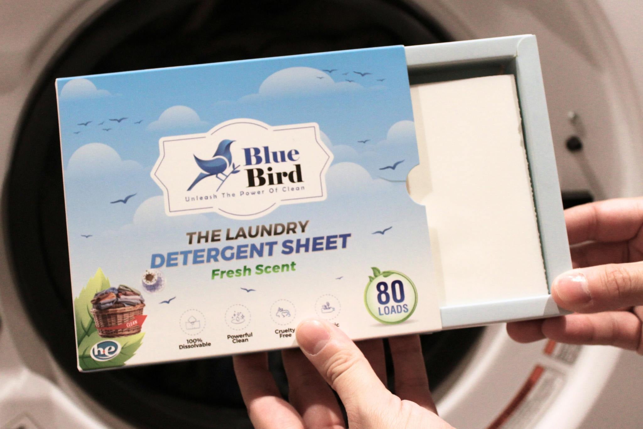 A person holds a Blue Bird laundry detergent sheet box in front of a washing machine door. The box is light blue with a sky-and-cloud design, and the product name reads 'The Laundry Detergent Sheet' in Fresh Scent.